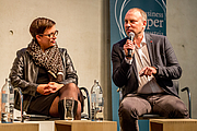 Ein Mann und eine Frau sitzen nebeneinander. Der Mann (rechts) spricht mit einem Mikrofon.